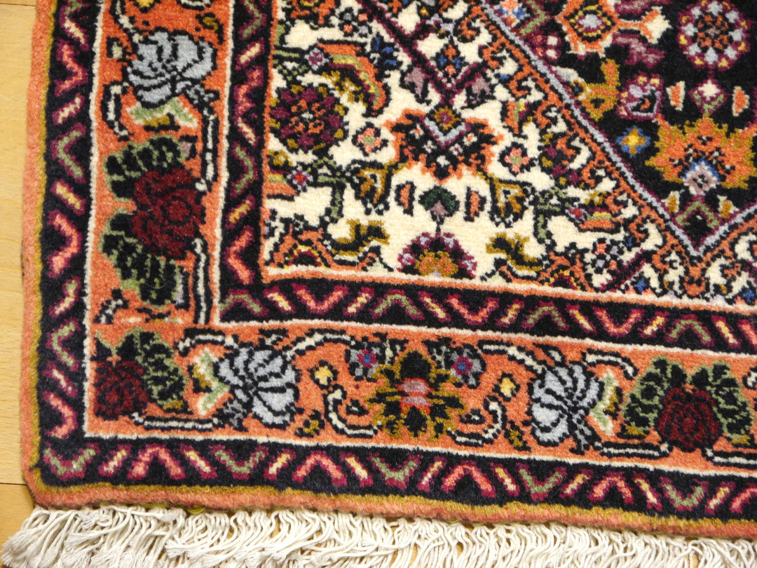 Ecke des Bidjar Tekab auf Holzboden: Terrakottabordüre mit graublauen und weinroten Rosen, marineblauer Zackenstreifen und Baumwollfransen
